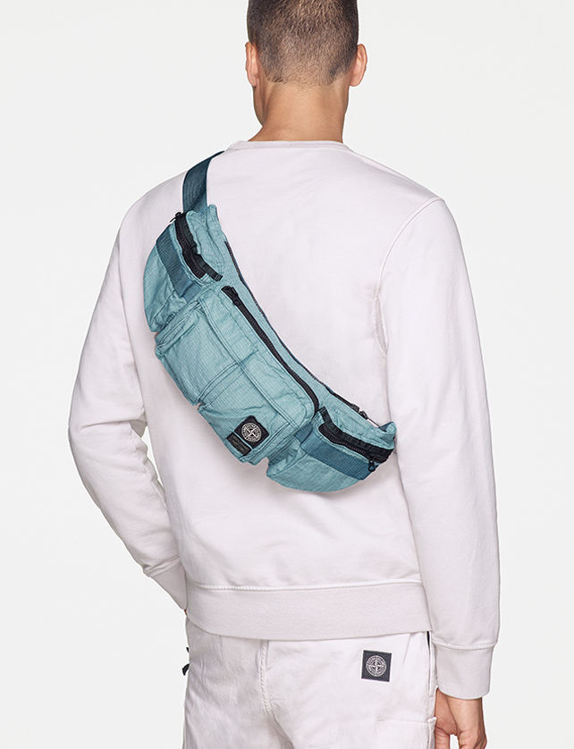 Back view of model wearing a white sweatshirt, white pants with the Stone Island patch on the back pocket and a light blue waist bag with black zippers and multiple pockets, worn over the shoulder.