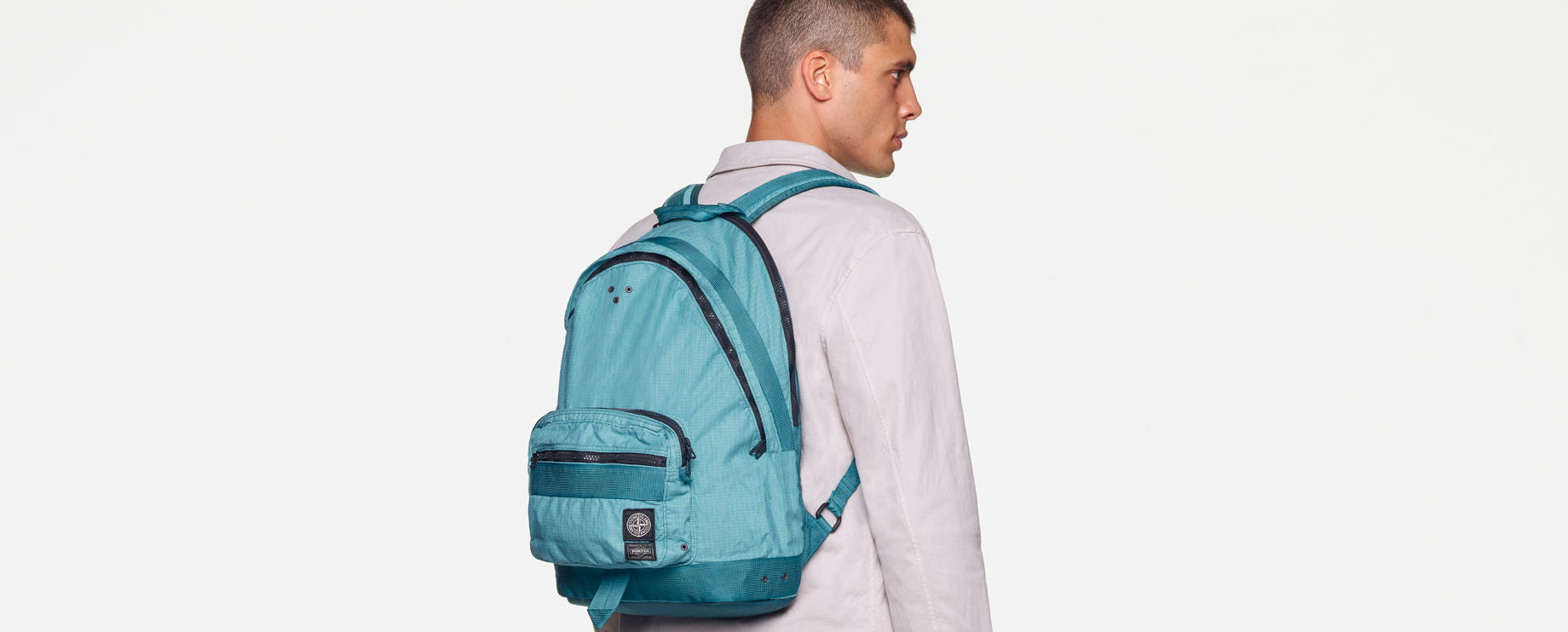 Back view of model wearing a white sweatshirt, white pants with the Stone Island patch on the back pocket and a light blue waist bag with black zippers and multiple pockets, worn over the shoulder.
