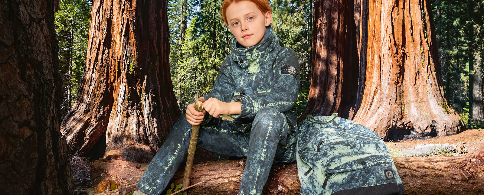 Junior model wearing blue and cream camouflage pants, a matching parka with hood, button fastening and a matching camouflage backpack with zipper fastening and patch pocket on front.