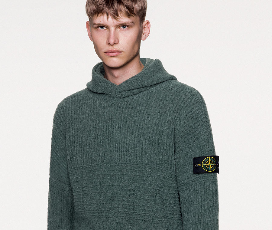Model wearing dark colored knit trousers with slanting hand pocket and a dark green hooded sweater with dropped shoulders, stitching motifs on the bust and sleeves and ribbed cuffs and bottom band.