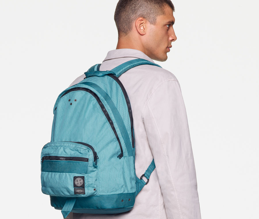 Back view of model wearing a white sweatshirt, white pants with the Stone Island patch on the back pocket and a light blue waist bag with black zippers and multiple pockets, worn over the shoulder.