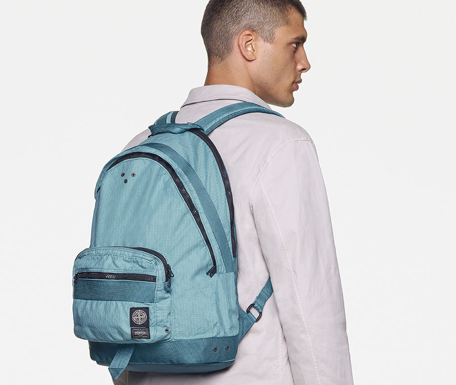 Back view of model wearing a white sweatshirt, white pants with the Stone Island patch on the back pocket and a light blue waist bag with black zippers and multiple pockets, worn over the shoulder.