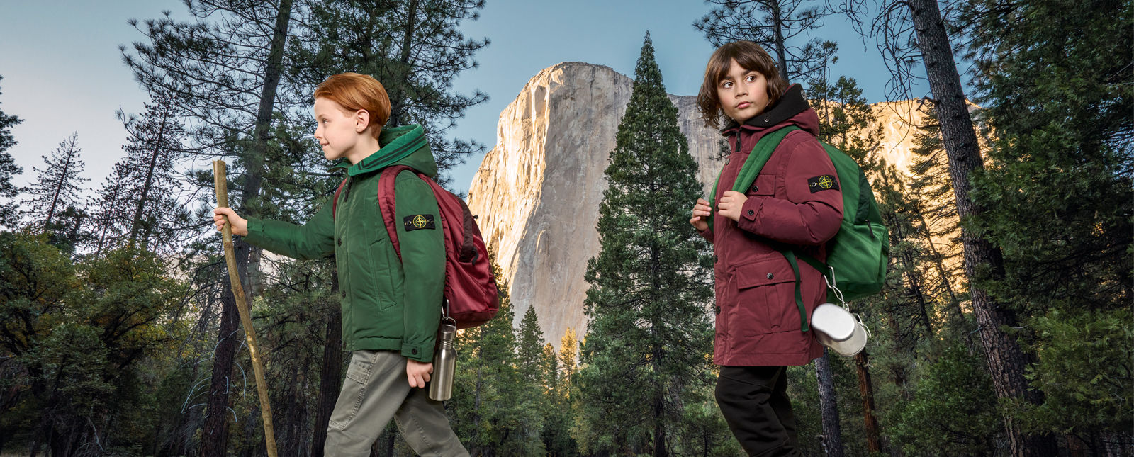 Two junior models against a desert backdrop, one wearing white pants, a red and white sweater and a red cap, the other one wearing a hooded red jacket with chest pockets and black zipper closure.