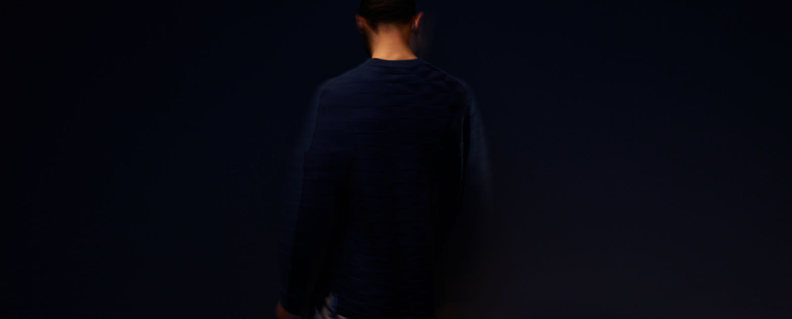 View of a model from the back wearing a dark blue crew neck sweater against a dark blue background