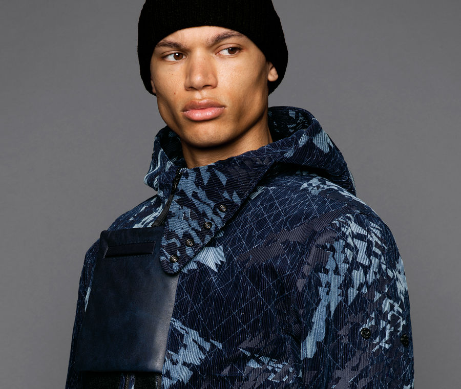 Cropped portrait of a model wearing short hooded jacket in an abstract pattern of dark and light blue hues with a square leather pocket on the chest