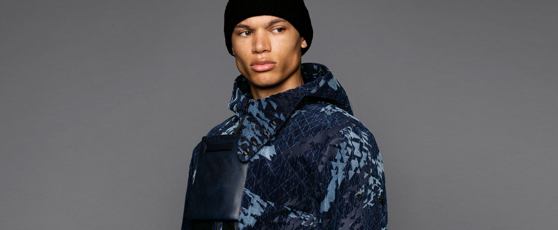 Cropped portrait of a model wearing short hooded jacket in an abstract pattern of dark and light blue hues with a square leather pocket on the chest