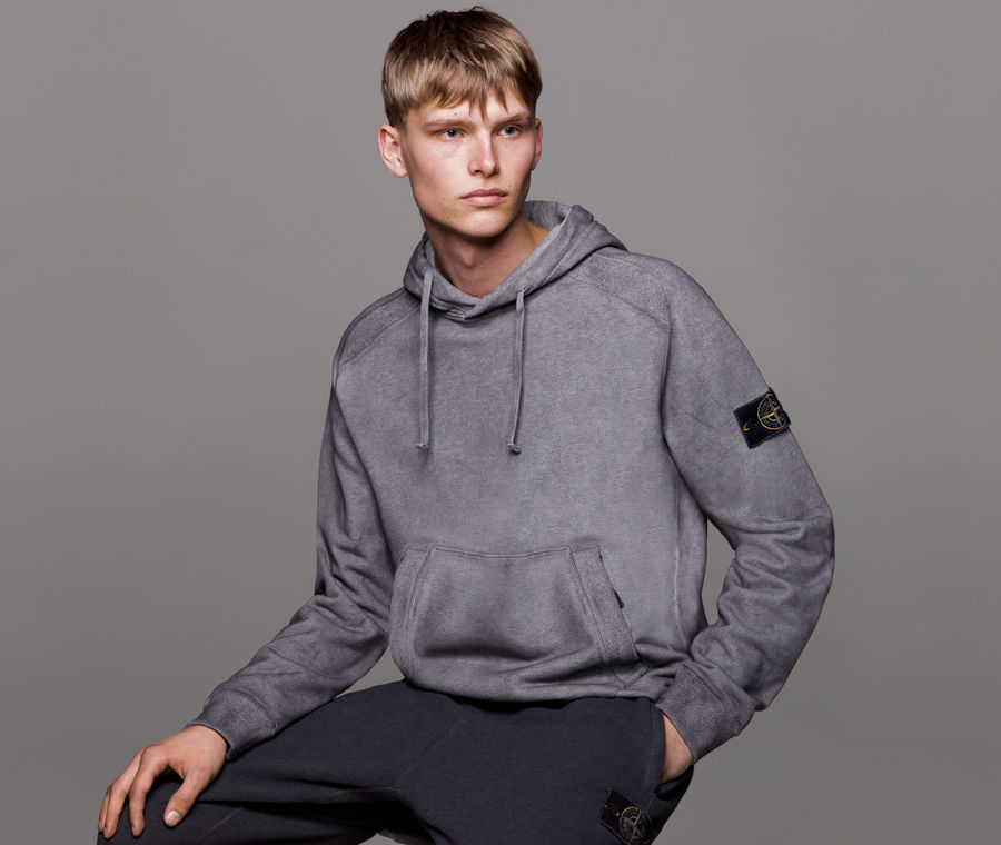 Model wearing grey hooded sweatshirt with drawstrings at the hood, kangaroo pocket, and Stone Island badge on the left sleeve.