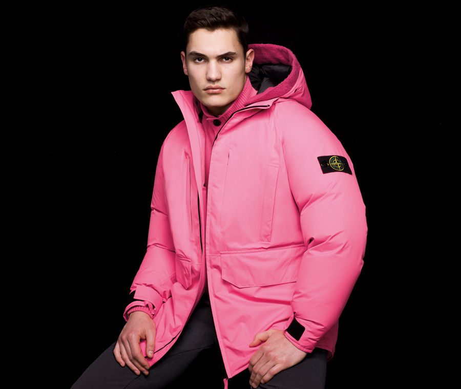 Side view of model wearing black pants and a pink hooded jacket with inner drawstring on back and the Stone Island badge on the upper left sleeve.