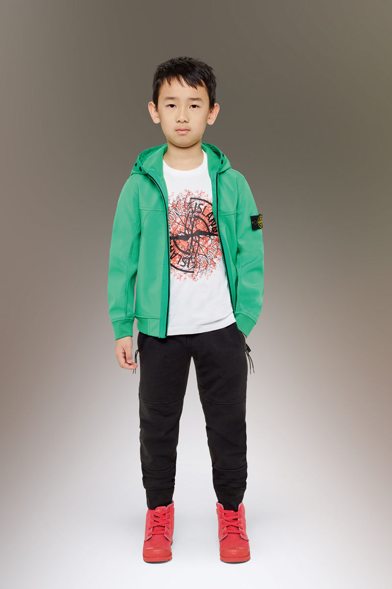 Junior model wearing red sneakers, black pants with slanting hand pockets, a white t shirt with a colorful print and the Stone Island compass logo, and a bright green sweatshirt with hood and zipper fastening.