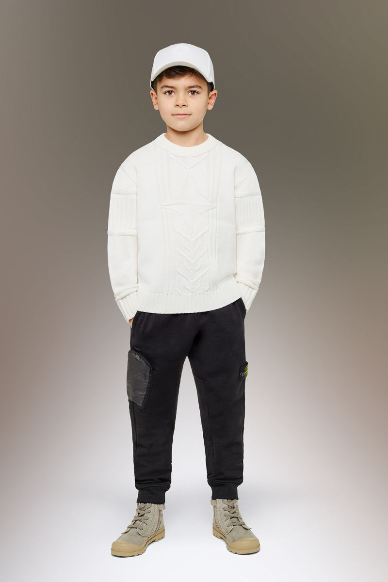Junior model wearing sand boots, black cargo pants with slanting hand pockets and the Stone Island badge on the left thigh, a white crewneck sweater and a white cap.