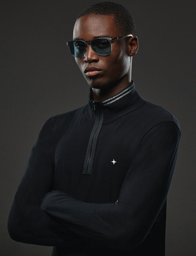 Upper body shot of a model wearing a pair of sunglasses with gray metal frame, blue drop shape lenses, a large metal bridge, adjustable nosepads and a black sweatshirt with high collar, half zipper fastening and the Stone Island star on the chest.