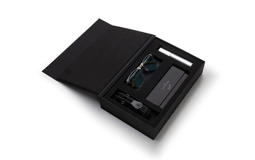 Still life image of an opened black rectangular box showing the items contained inside: a pair of metal frame glasses, a toggle with the Stone Island logo, a metal cylinder and a case with both the Stone Island and the Persol logos on the flap.