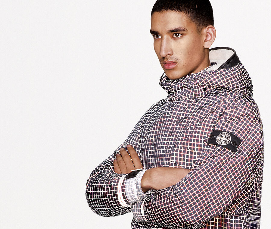 Portrait shot of model with folded arms wearing a black check print jacket with hood, light gray colored cuffs and the black and white Stone Island research badge on the upper left arm.