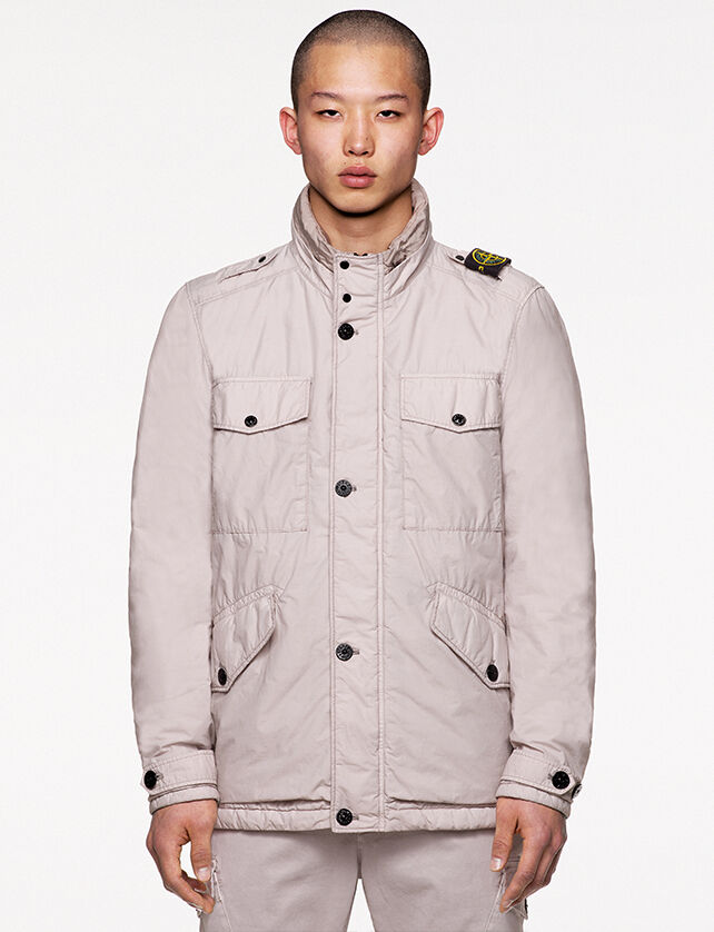 Model wearing off white pants and a matching field jacket with standing collar with two snap fastening, chest and hand pockets with flap and snap fastening and the Stone Island badge on the left shoulder.