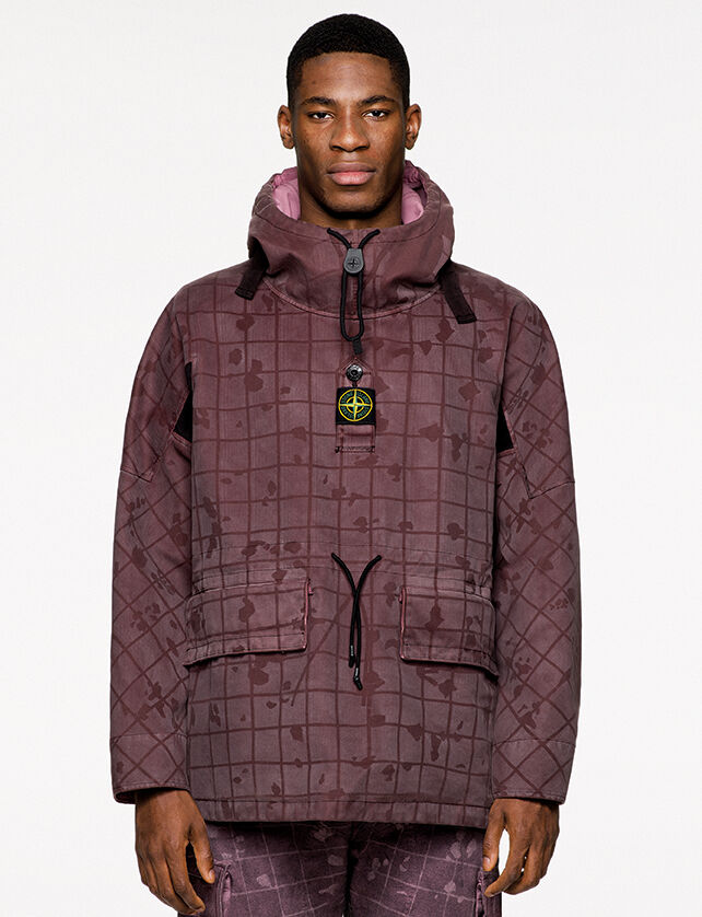 Model wearing burgundy check and paint splatter print pants and a matching anorak with standing collar, front hand pockets with flap and central buttoned strap bearing the Stone Island badge.
