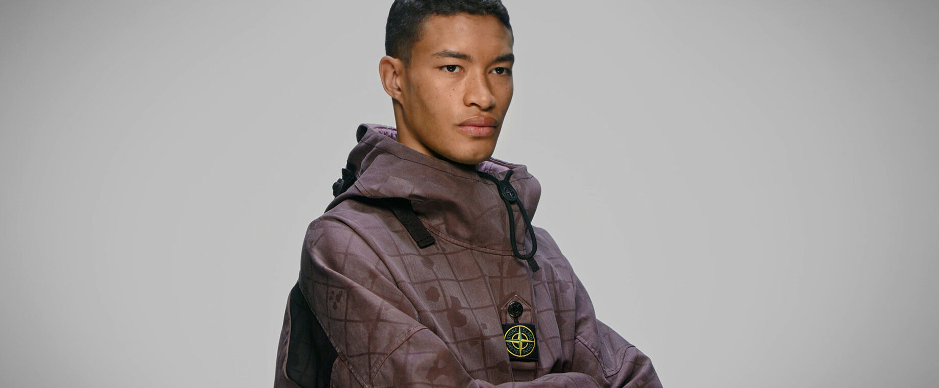 Close up of model wearing burgundy check and paint splatter print anorak with standing collar, toggle and central buttoned strap bearing the Stone Island badge.