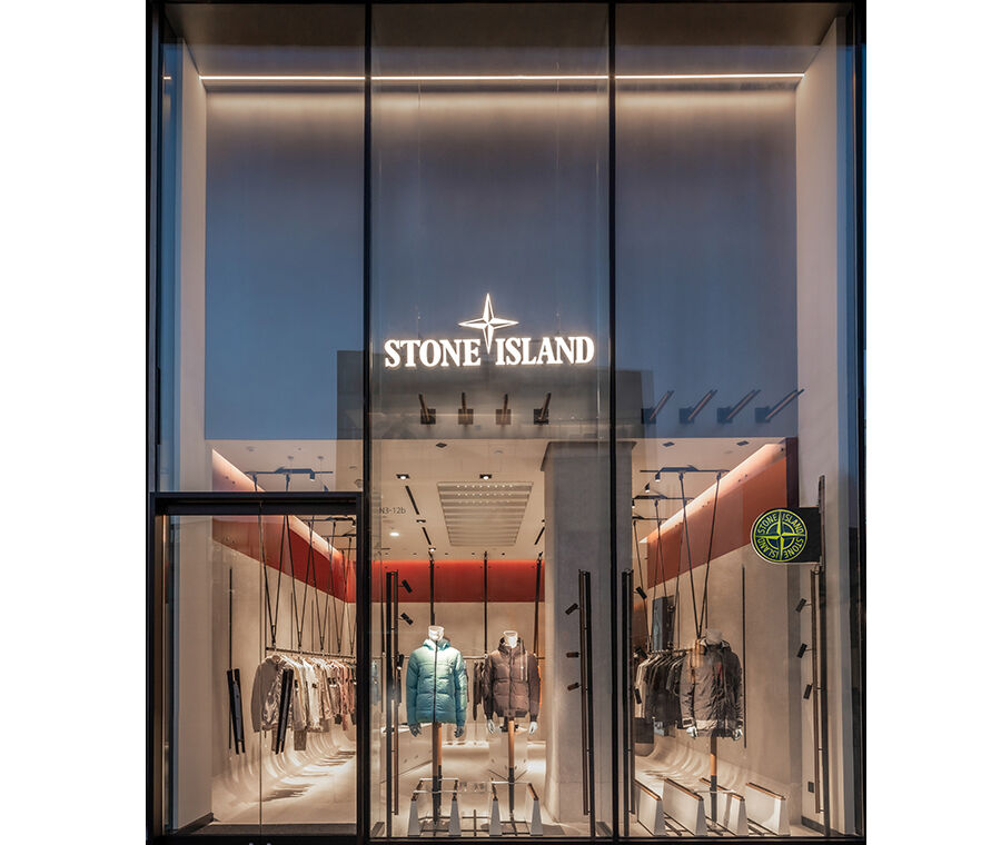 Interior view of a store with red accents, garments arranged by color hanging on racks along the side walls and two white display tables with shoes and accessories in the center.