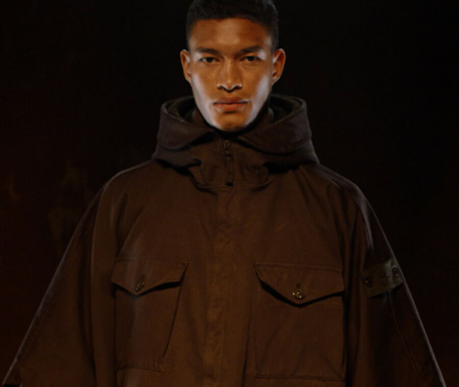 Close up shot of model wearing dark brown hooded jacket with hidden zipper closure, two bellows chest pockets with flap and button fastening and the Stone Island ghost badge on upper left arm.