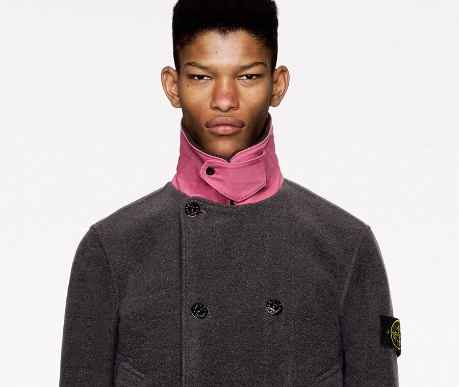 Model wearing dark colored pants and a dark gray pea coat with pink standing collar, chin strap held by button fastening, double breasted buttoning, two patch pockets and pink parallel stripes on the cuffs and lower hem.