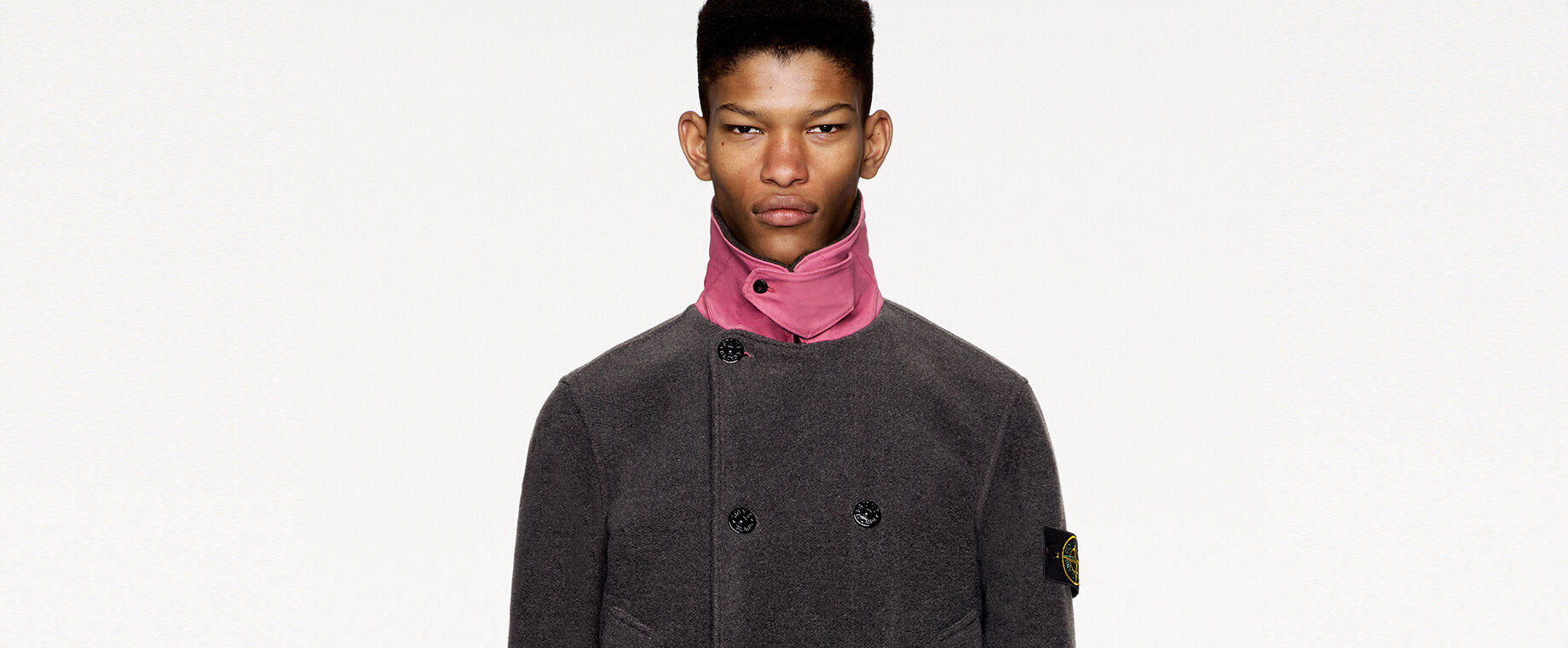 Model wearing dark colored pants and a dark gray pea coat with pink standing collar, chin strap held by button fastening, double breasted buttoning, two patch pockets and pink parallel stripes on the cuffs and lower hem.