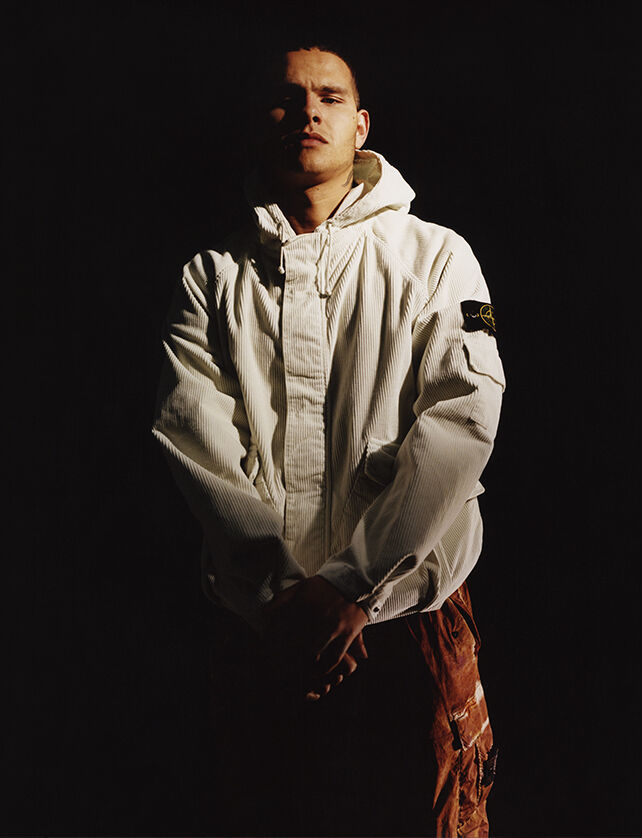 A model wearing orange camouflage cargo pants and an off white hooded jacket with the Stone Island badge on the upper left arm.