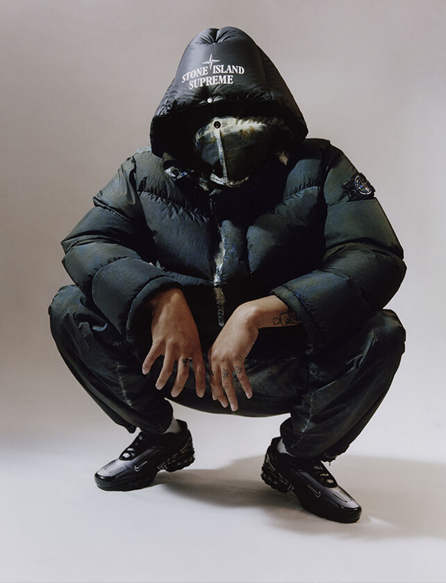 One crouching model wearing dark colored cargo pants with white edged patch bellows pocket on the left thigh and a matching down jacket with face flap and a hood with the Stone Island Supreme logo.