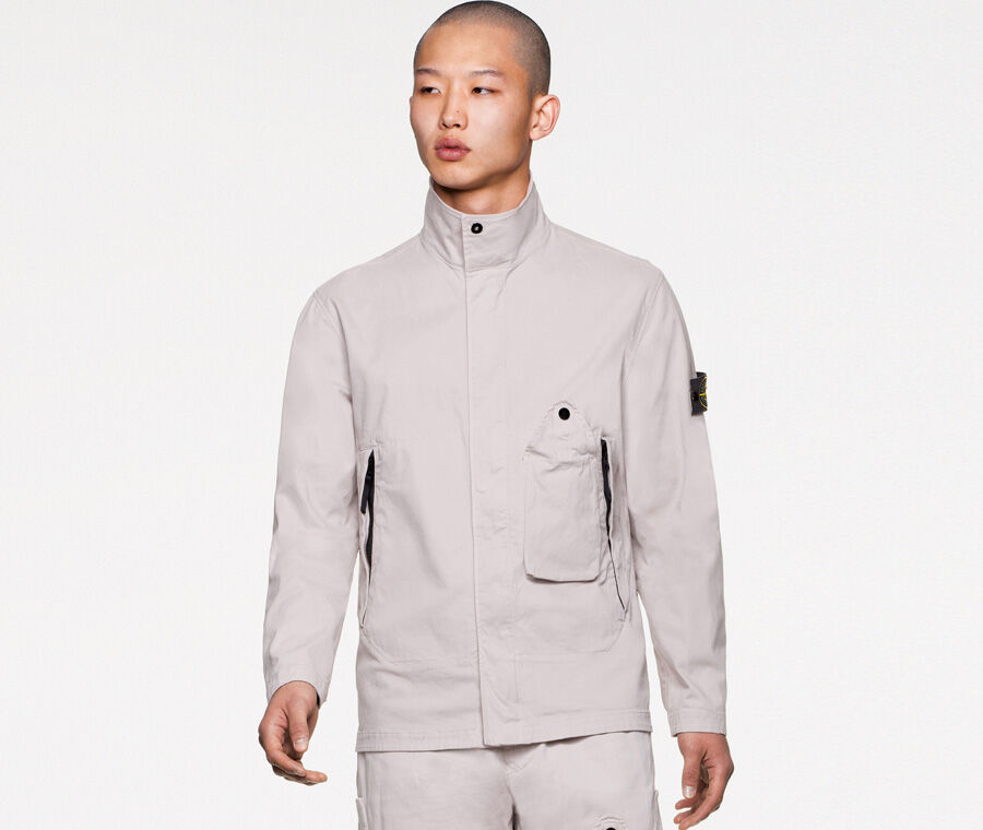 Model wearing off white pants and a matching overshirt featuring a standing collar with small button fastening, slanting hand pockets with zipper and one front bellows patch pocket with a standing triangular flap fastened by a snap.