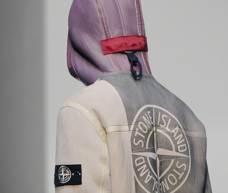 Close up shot of model seen from the back, wearing a gray, off white and purple suede sheepskin jacket with the Stone Island logo printed on the back and a hood featuring parallel vertical drawstrings with black toggle and pull.