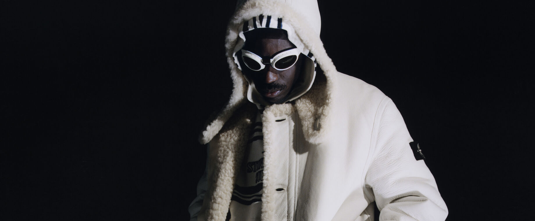 Against a black background, model wearing a white hooded sheepskin jacket, a black and white striped hoodie and white goggles with dark lenses.