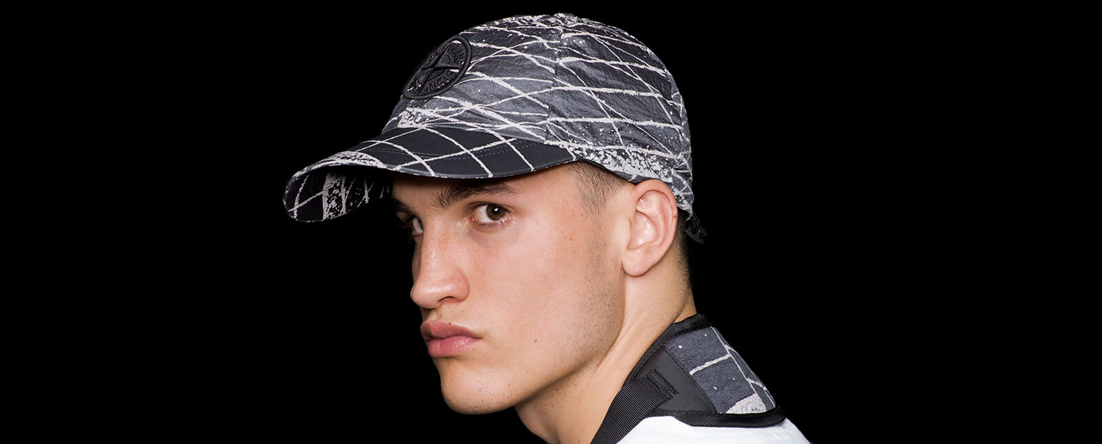 Close up shot of model wearing a dark gray and white print cap with the Stone Island Compass logo embroidery on the front and an elasticated drawstring on the back.