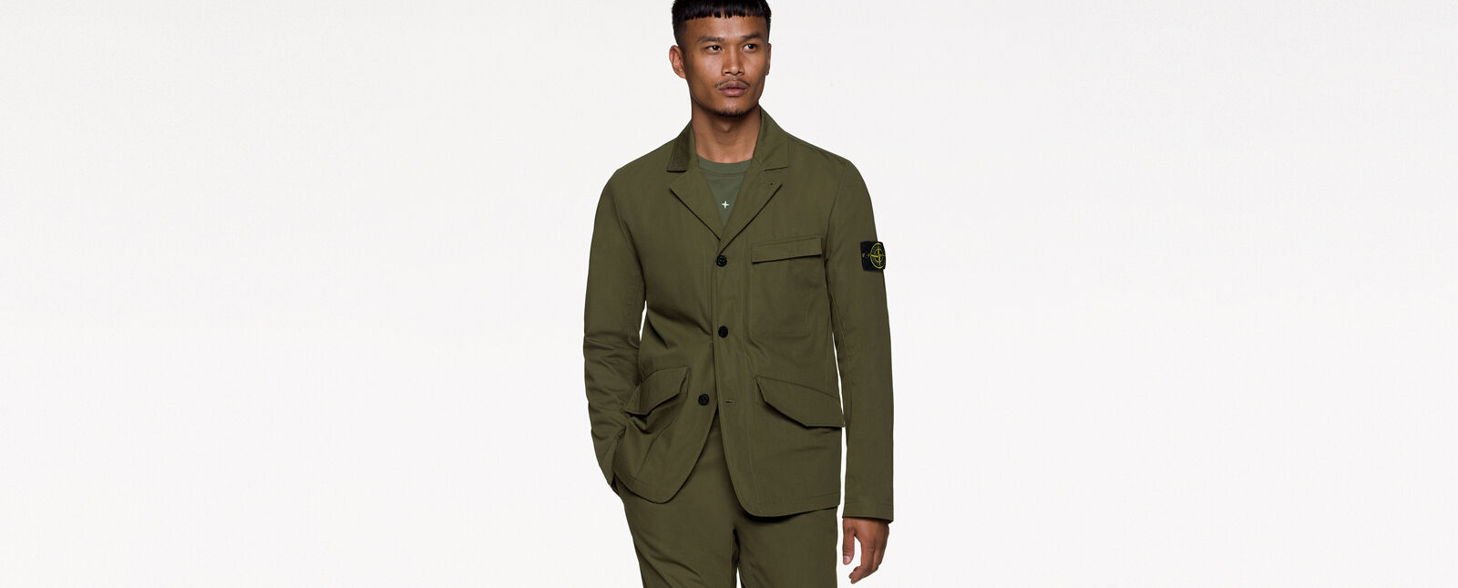 Model wearing military green pants and a matching jacket with collar, three button fastening, a chest pocket with flap, two hand pockets with slanting flap and the Stone Island badge on the upper left sleeve.