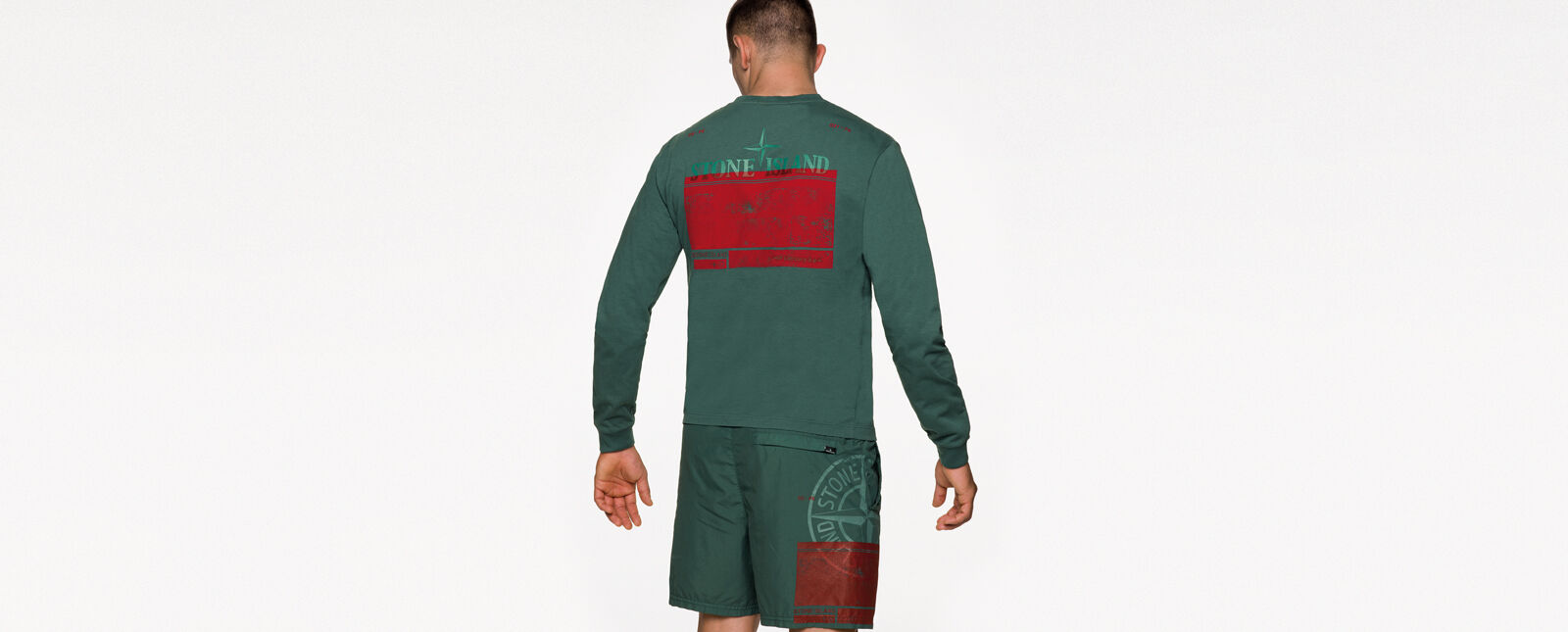 Back view of model wearing a green crewneck t shirt with long sleeves and a red print on the back and green shorts with a red print and the Stone Island compass rose logo on the right thigh.