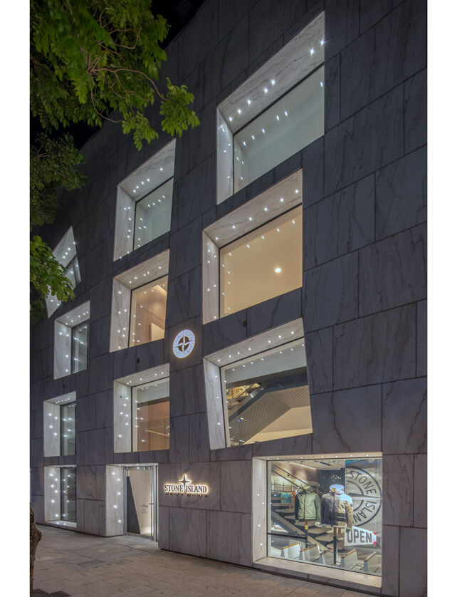 Exterior view of a lit up store with a tree in front of it, seven irregularly placed windows, the entrance on the left, a back lit Stone Island logo and compass rose in the middle.