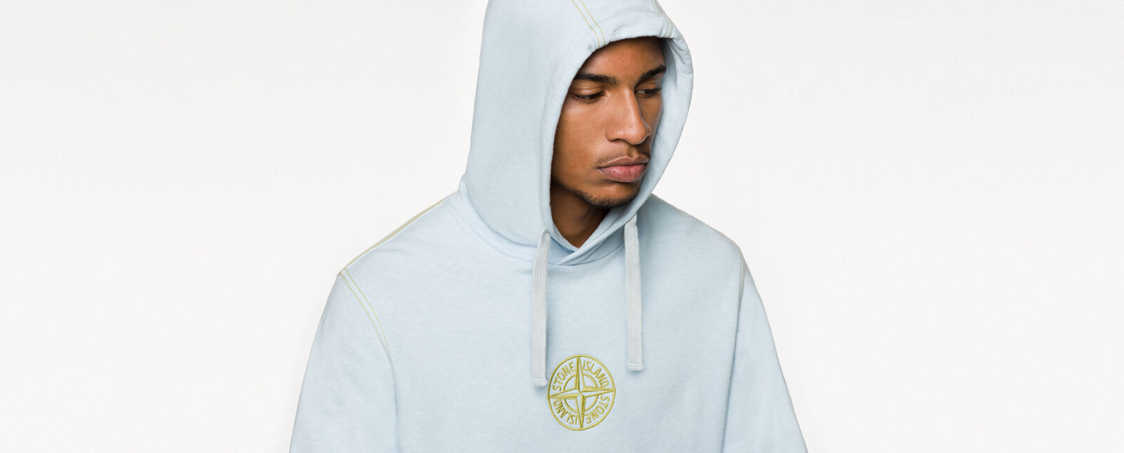 Close up of model wearing a light blue sweatshirt with hood, yellow stitching and the Stone Island compass rose logo embroidered in yellow on the chest.