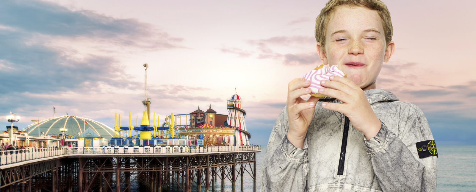 View of a pier amusement park and a waterfront promenade on the left and a close up shot of a junior model eating a donut, wearing an off white dust effect hooded jacket with half zipper and the Stone Island badge on the upper left arm.