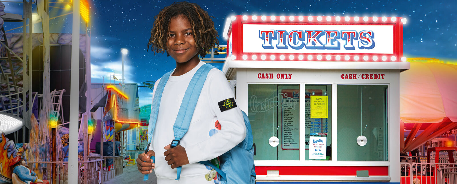 Amusement park rides and ticket office in the background and a close up shot of a junior model wearing a white crewneck sweatshirt and a light blue backpack with adjustable straps.