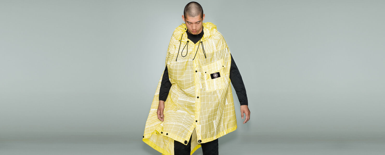 Model wearing black pants, a matching crewneck top and a yellow cape with a white print, a hood with drawstrings, button fastening and a patch pocket with flap, snaps and the Stone Island research badge.