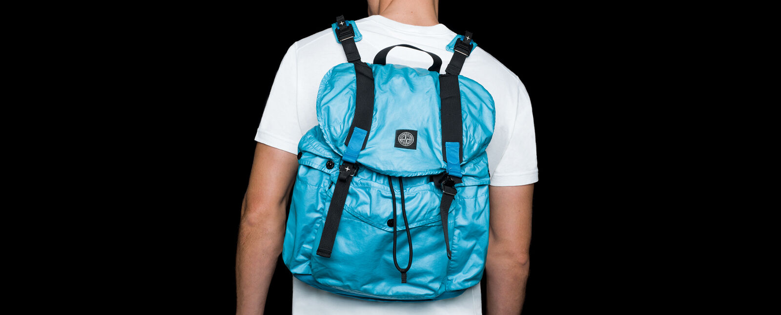 Back view of model wearing a white crewneck t shirt and a light blue shiny effect backpack with shoulder straps, flap cover with double fastening and a central pocket with flap and snap.