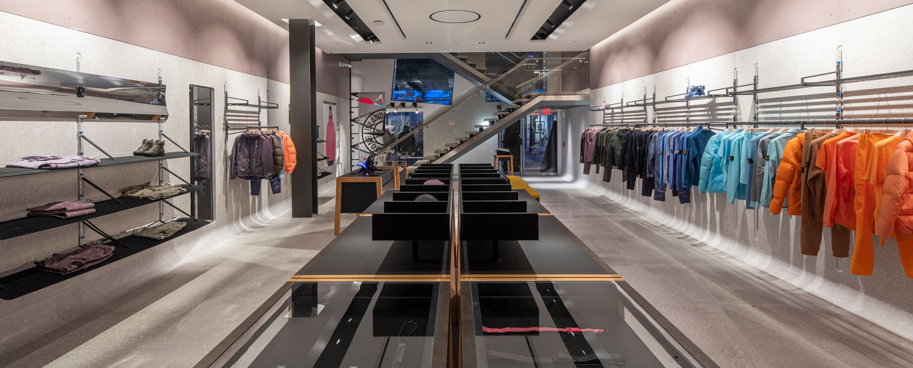 Interior view of a store with off white walls, floor and ceiling, garments arranged by color hanging on a rack on the right, a display table with accessories in the center and on the left racks with a pair of shoes, garments both hanging and folded.