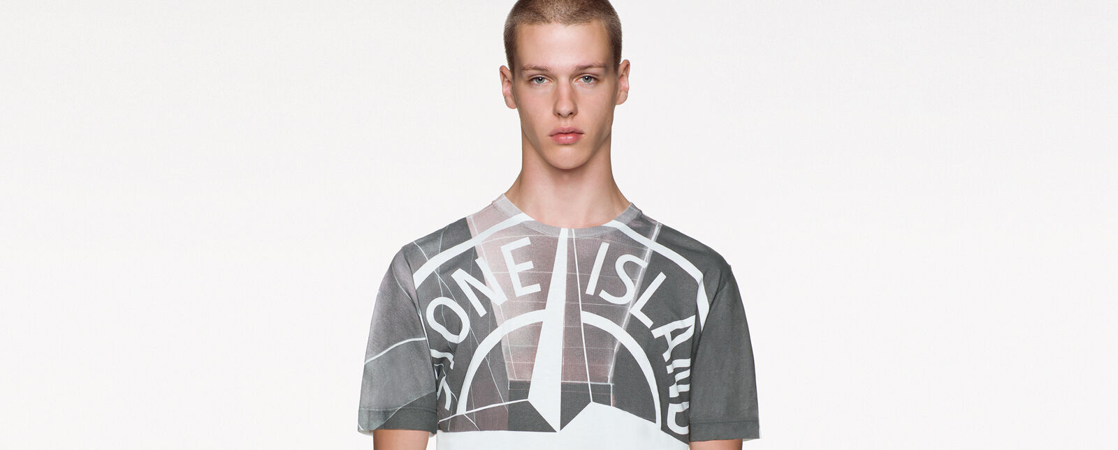 Close up shot of model wearing a gray print crewneck t shirt with short sleeves and a large white Stone Island compass rose logo print on the bust.