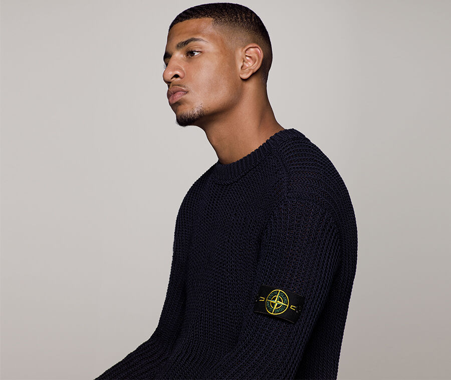 Close up shot of model wearing a dark colored sweater with a ribbed crewneck, and the Stone Island badge on the upper left sleeve.