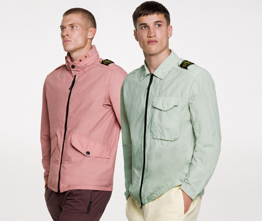 Two models wearing the same outfit, consisting of dark green shorts and a matching overshirt with two way zipper fastening, a bellows pocket with flap and snaps on the chest and the Stone Island badge on the upper left arm.