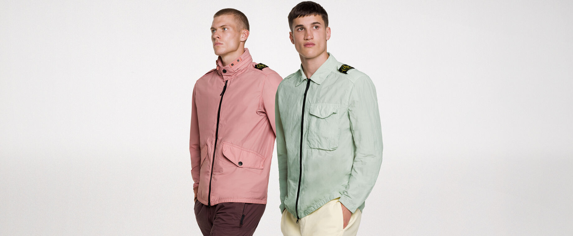 Two models wearing the same outfit, consisting of dark green shorts and a matching overshirt with two way zipper fastening, a bellows pocket with flap and snaps on the chest and the Stone Island badge on the upper left arm.