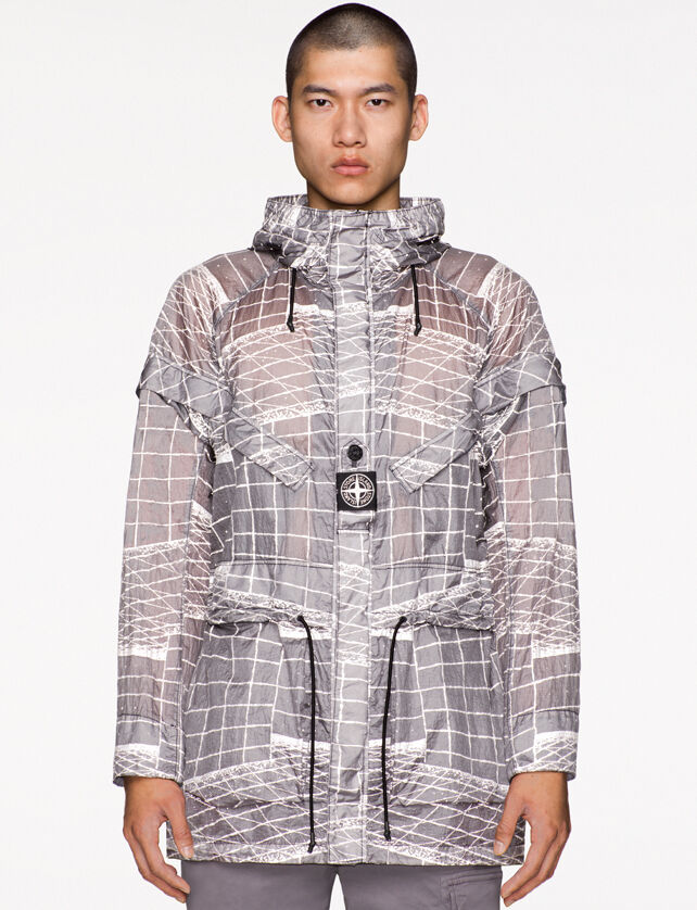 Model wearing gray pants and a light gray see through hooded jacket with a white print, drawstring at waist and a fastening placket with a buttoned central strap on the chest featuring the Stone Island research badge.