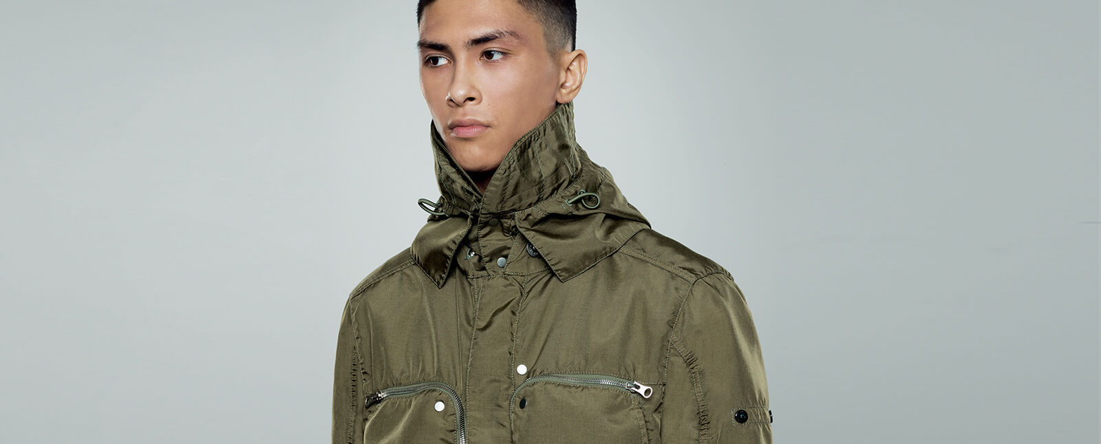 Close up shot of model wearing a military green jacket with standing collar with drawstring and two chest pockets with zipper fastening.
