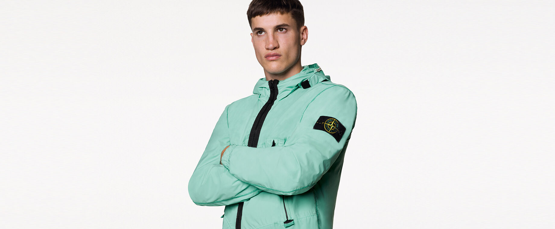 Model wearing a mint green light jacket with hood, black zipper fastening and the Stone Island badge on the upper left sleeve.