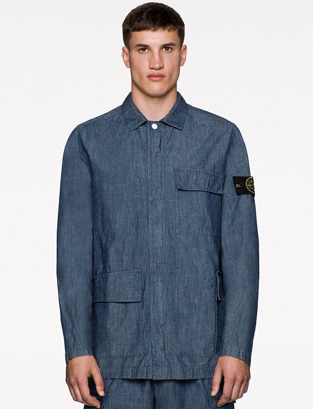 Model wearing indigo chambray canvas pants and a matching overshirt with collar, one visible button, fastening hidden under a flap and two patch pockets with flap, one at left chest and one on the right hip.