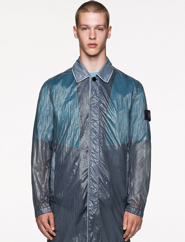 Model wearing a gray blue shiny effect trench coat with a collar with snap, hidden fastening, standing hand pockets and the Stone Island badge on the upper left arm.