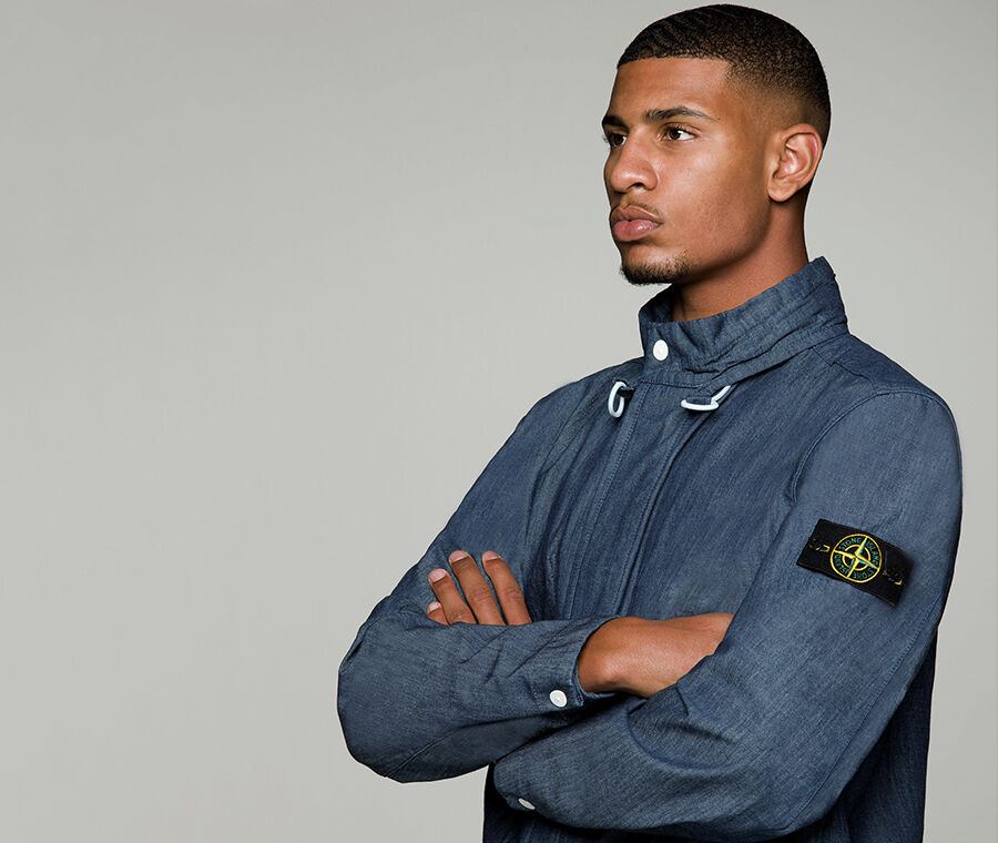 Close up shot of model wearing indigo chambray canvas jacket with button up collar, white drawstring, fastening hidden under a flap, cuffs with white snap fastening and the Stone Island badge on the upper left sleeve.