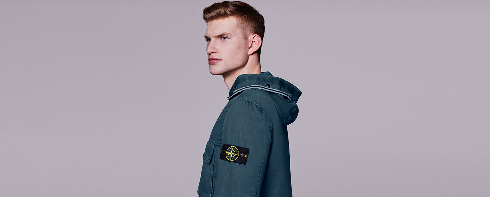 Side view of model wearing a dark blue overshirt with a chest pocket with flap, a hood edged with two parallel light blue strips and the Stone Island badge on the upper left sleeve.